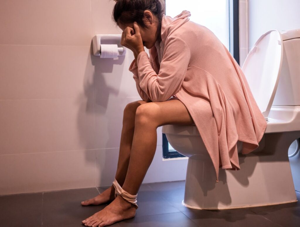 Mulher sentada no vaso sanitário com aparente desconforto que pode ser causado por fissura anal.