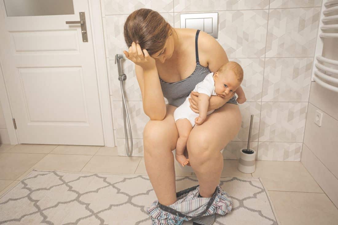Mulher com bebê no colo sofrendo dor intensa causada por fissura anal crônica, ilustrando desconforto durante a evacuação.