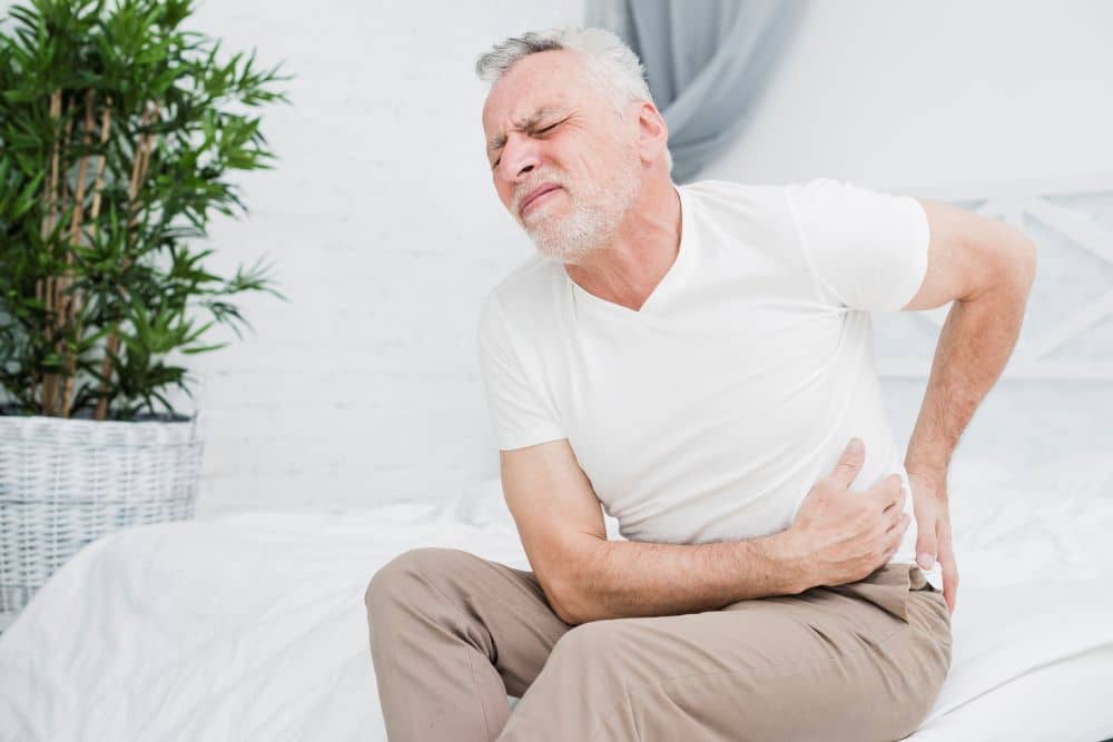 Homem de meia-idade sentado no sofá com expressão de dor segurando as costas, representando desconforto causado por problemas renais crônicos.