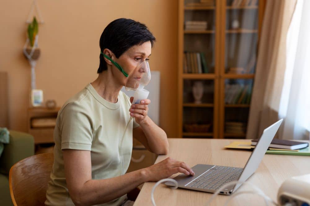A imagem retrata uma mulher adulta em ambiente domiciliar, sentada à mesa em frente a um computador, utilizando uma máscara de nebulização ou dispositivo de oxigenoterapia, o que sugere o manejo de uma condição respiratória crônica. A cena remete ao acompanhamento de pacientes com doenças pulmonares, como a Doença Pulmonar Obstrutiva Crônica (DPOC), especialmente em contextos de tratamento contínuo e monitoramento fora do ambiente hospitalar. O uso do dispositivo respiratório evidencia a importância do controle dos sintomas, como dispneia e dificuldade ventilatória, comuns na DPOC, além de reforçar o papel da terapia inalatória e, em casos mais avançados, do suporte com oxigênio domiciliar. A presença do computador sugere a utilização de recursos de telemedicina ou prontuários eletrônicos, destacando a relevância do acompanhamento médico remoto, da adesão ao tratamento e da orientação contínua por profissionais de saúde. O ambiente acolhedor e doméstico reforça a necessidade de uma abordagem integral no cuidado do paciente com DPOC, que inclui não apenas o uso correto de medicamentos broncodilatadores e corticoides inalados, mas também a educação em saúde, a reabilitação pulmonar, o acompanhamento regular e medidas de cessação do tabagismo. A imagem ilustra, portanto, o cuidado centrado no paciente e a importância do tratamento individualizado para a melhoria da qualidade de vida e redução das exacerbações associadas à DPOC.