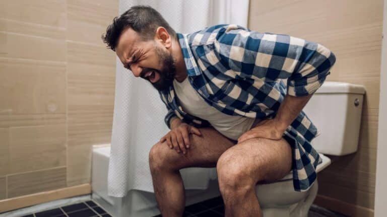 Homem com expressão de dor sentado no vaso sanitário, ilustrando desconforto e possível sangramento ao evacuar.